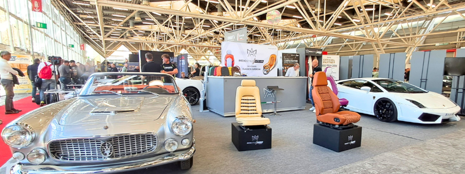 stand fieristico con esposizione a sinistra di un'auto d'epoca, al centro 3 sedili con diverse lavorazioni e a destra un'auto sportiva